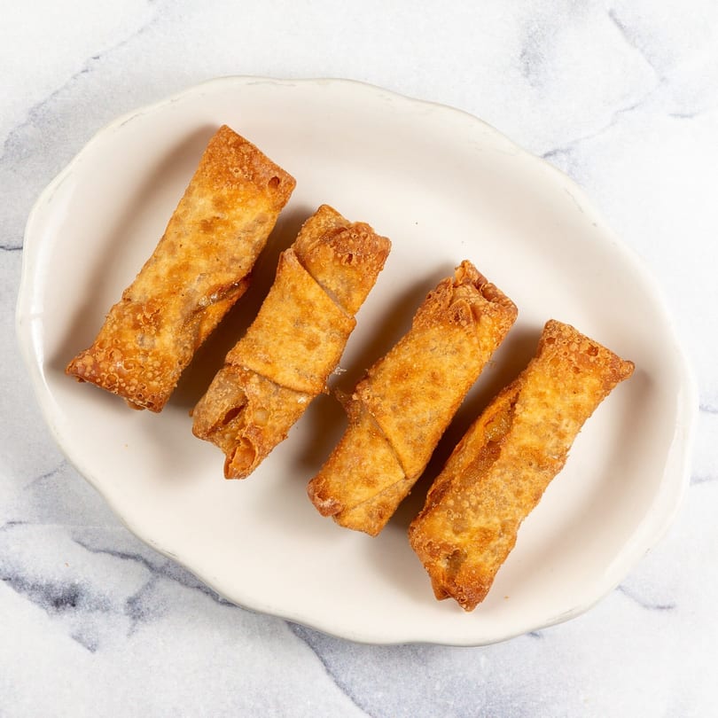 Four golden-brown fried egg rolls arranged on a white, irregularly shaped plate against a marble background.