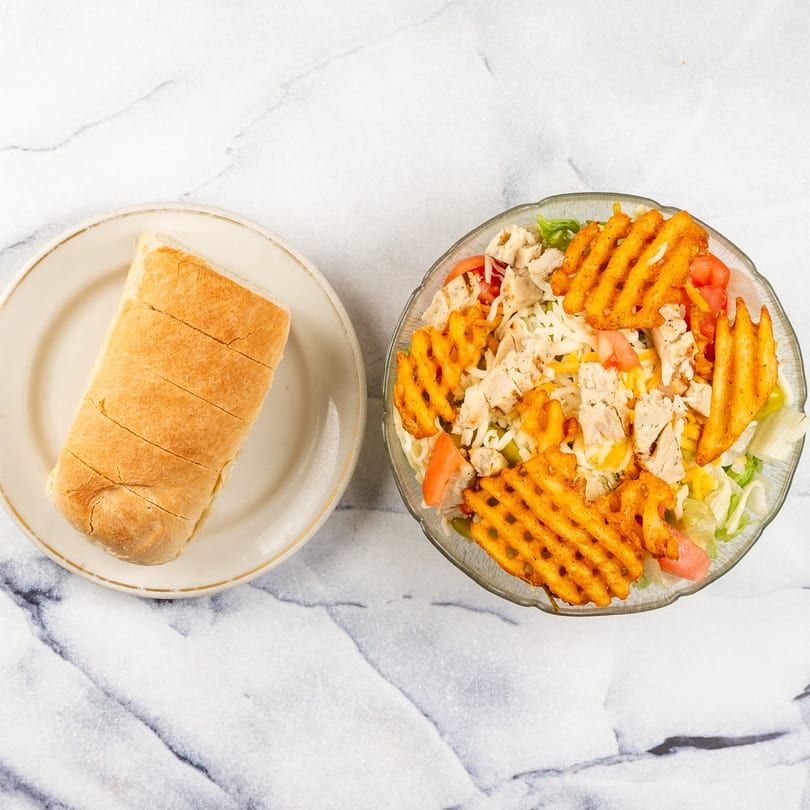 A sliced sub roll on a plate next to a salad with chicken, cheese, tomatoes, and waffle fries.