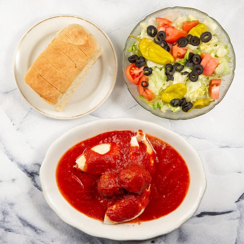 Stuffed shells with meatballs in red sauce, a side salad, and sliced bread on a marble surface.