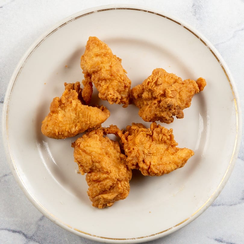 Five golden-brown fried chicken tenders on a white plate with a gold rim.